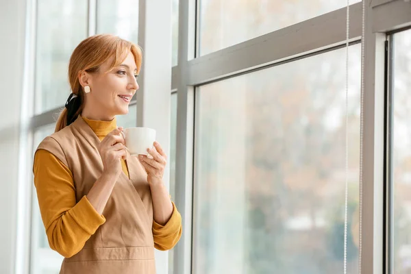 Beautiful mature woman drinking coffee near window in office — Stock Photo, Image