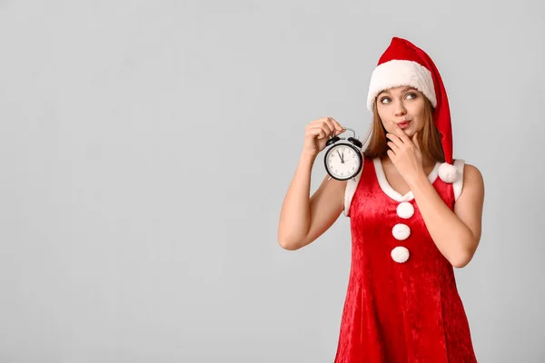 Bedachtzame jonge vrouw in Santa kostuum en met wekker op lichte achtergrond. Kerst countdown concept — Stockfoto