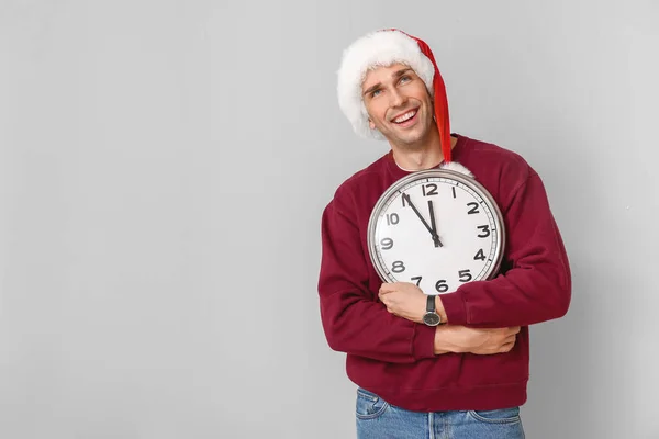 Young man in Santa hat and with clock on light background. Christmas countdown concept — Stock Fotó