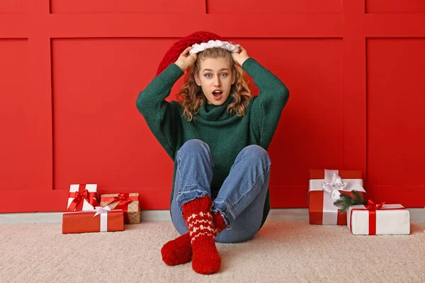 Překvapená mladá žena v zimním oblečení a s vánočními dárky v blízkosti barevné stěny — Stock fotografie