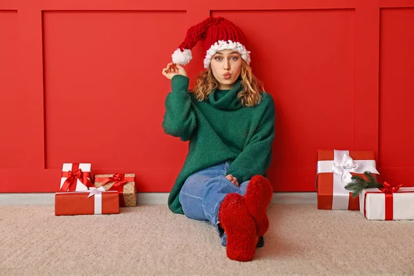 Happy young woman in winter clothes and with Christmas gifts sitting near color wall — Stock Photo, Image