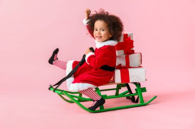Noel Baba kostümlü küçük Afro-Amerikalı kız, kızakları ve renk arka planında hediye kutuları var.