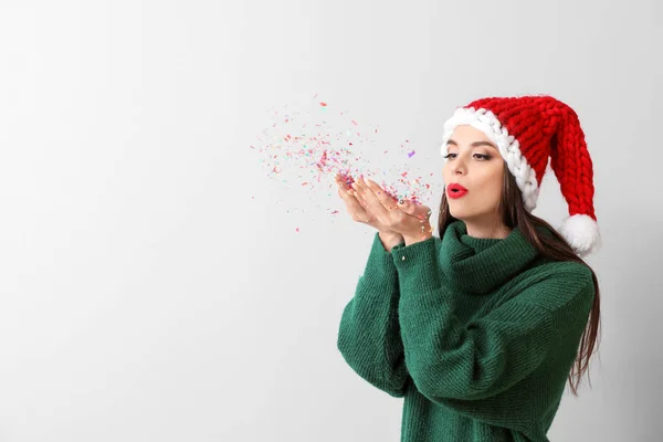 Bella giovane donna in cappello Babbo Natale e con coriandoli su sfondo chiaro — Foto Stock