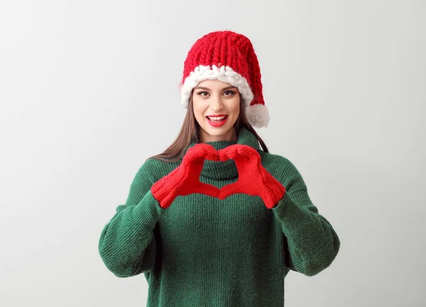 Молодая женщина в шляпе Санта-Клауса делает сердце руками на светлом фоне — стоковое фото