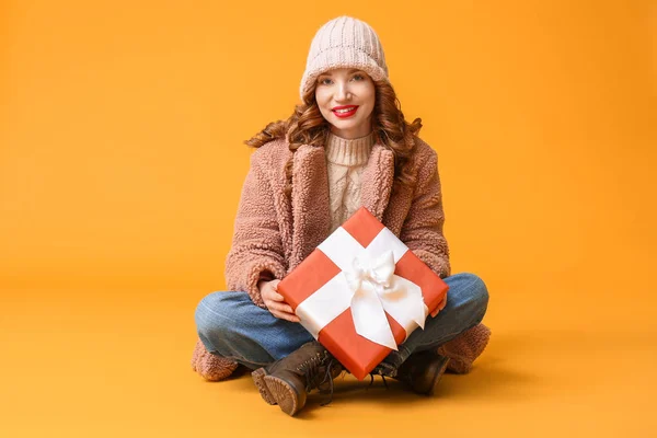 Happy young woman in winter clothes and with Christmas gift on color background — Stock Photo, Image
