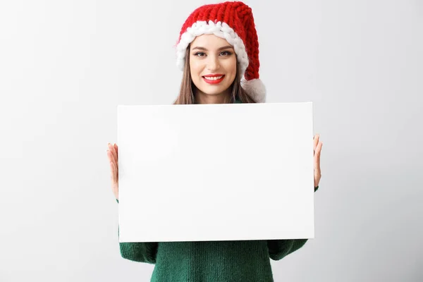 Piękna młoda kobieta w kapeluszu Santa i z pustym plakatem na jasnym tle — Zdjęcie stockowe