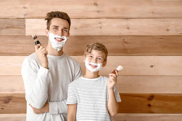 Padre e il suo piccolo figlio con schiuma da barba sul viso e pennelli sullo sfondo di legno — Foto Stock
