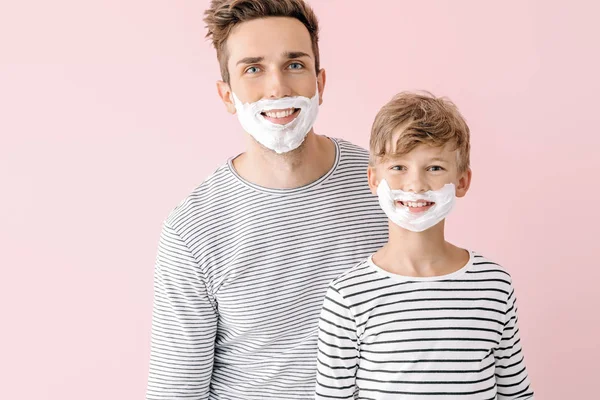 Father and his little son with shaving foam on their faces against color background