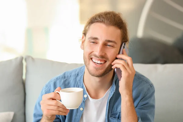 Handsome man with cup of coffee talking by mobile phone at home — Stock Photo, Image