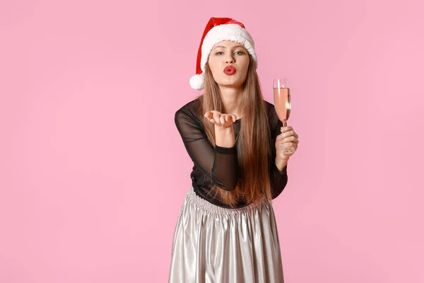 Happy young woman in Santa Claus hat and with glass of champagne blowing kiss on color background — Stock Photo, Image