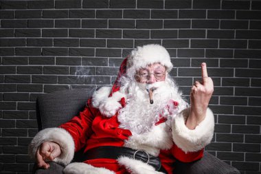 Sigara içen komik sarhoş Noel Baba orta parmağını tuğla arka planda koltukta otururken gösteriyor.