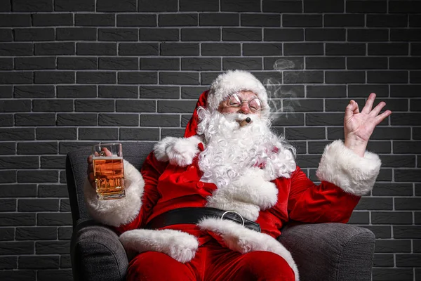 Zabawny pijany Święty Mikołaj pokazuje Ok siedząc w fotelu na tle cegły — Zdjęcie stockowe