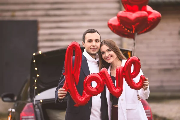 Feliz pareja joven con globo de aire en forma de palabra AMOR cerca de coche al aire libre. Celebración de San Valentín —  Fotos de Stock