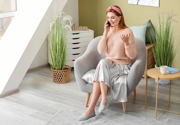 Beautiful woman talking by phone while sitting in armchair at home — Stock Photo, Image