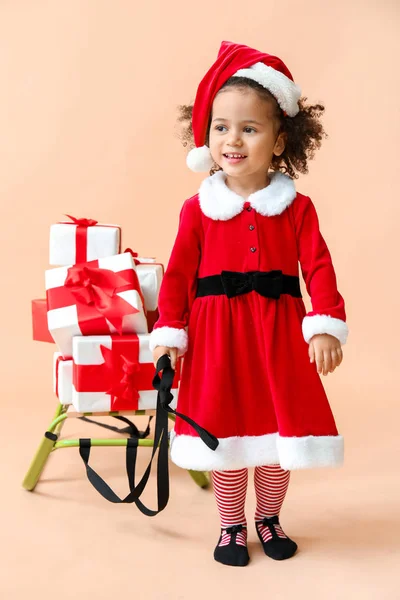 Petite fille afro-américaine en costume de Père Noël, avec des cadeaux et des traîneaux sur fond de couleur — Photo