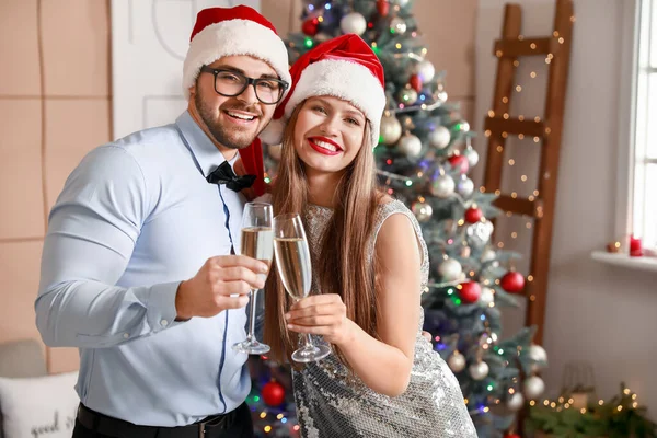 Happy young couple in Santa Claus hats and with glasses of champagne at home — Stock Photo, Image
