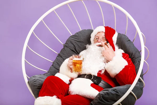 Funny Santa Claus drinking alcohol while sitting in armchair on color background — Stock Photo, Image