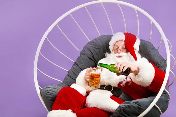 Funny Santa Claus drinking alcohol while sitting in armchair on color background — Stock Photo, Image