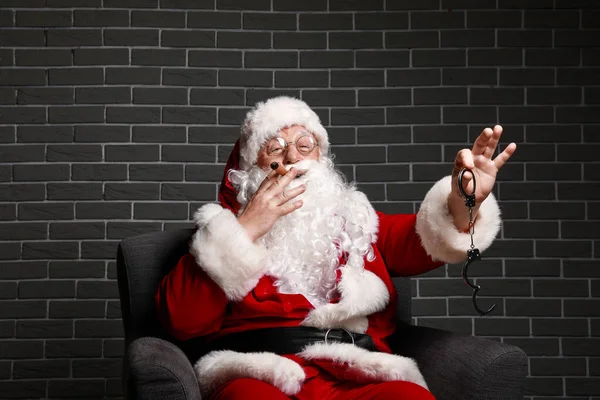 Funny drunk Santa Claus with handcuffs sitting in armchair against brick background — Stock Photo, Image