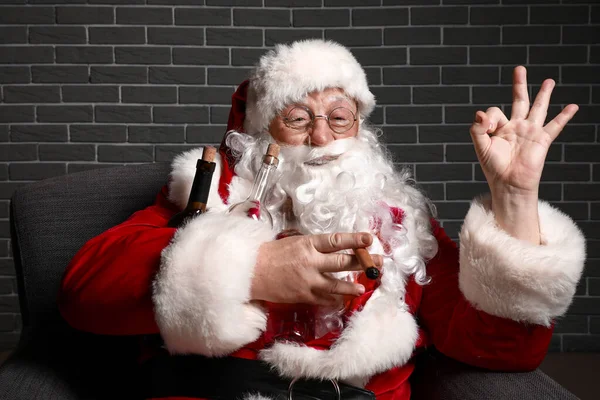 Zabawny pijany Święty Mikołaj pokazuje Ok siedząc w fotelu na tle cegły — Zdjęcie stockowe
