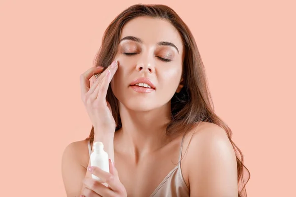Beautiful young woman applying cream onto her skin against color background