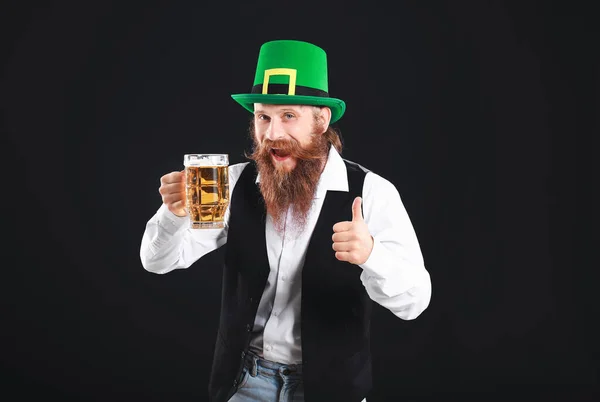 Hombre barbudo con un vaso de cerveza mostrando un gesto de pulgar hacia arriba sobre un fondo oscuro. Celebración del Día de San Patricio — Foto de Stock
