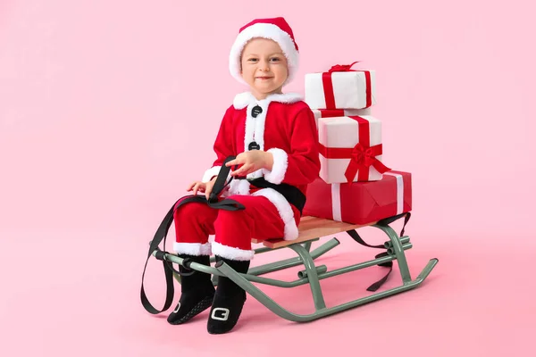 Niedlicher kleiner Junge im Weihnachtsmann-Kostüm, mit Schlitten und Weihnachtsgeschenken auf farbigem Hintergrund — Stockfoto