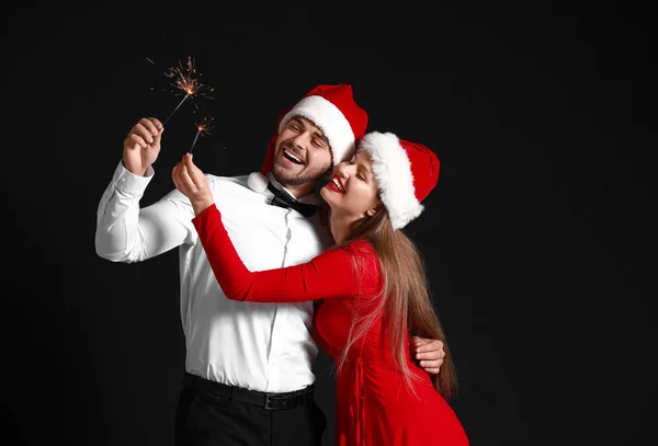 Happy couple in Santa Claus hats and with Christmas sparklers on dark background — Stock Photo, Image