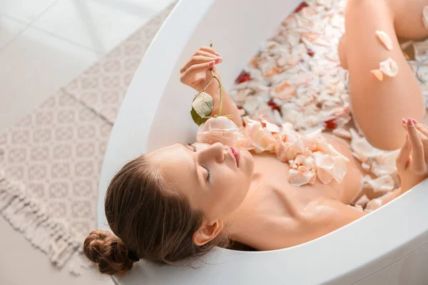 Beautiful young woman taking bath with flower petals at home — Stock Photo, Image