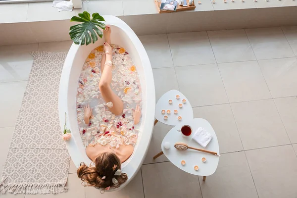 Hermosa joven tomando baño con pétalos de flores en casa — Foto de Stock