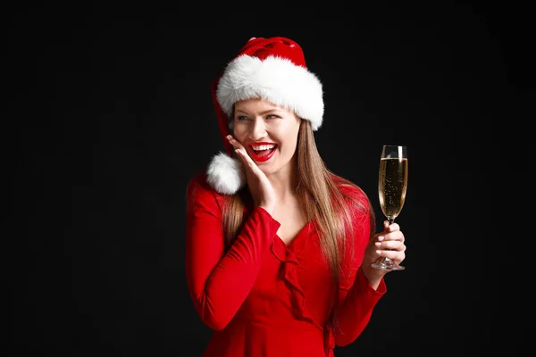 Happy young woman in Santa Claus hat and with glass of champagne on dark background — Stock Photo, Image