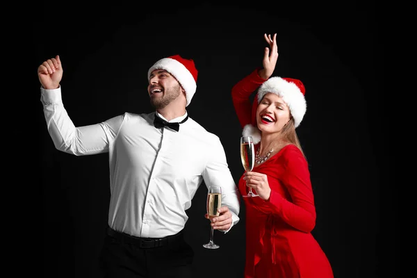 Casal feliz em chapéus de Papai Noel e com copos de champanhe no fundo escuro — Fotografia de Stock