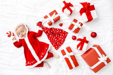 Noel Baba kostümü içinde küçük şirin bir bebek ve yatağında hediyeler var.