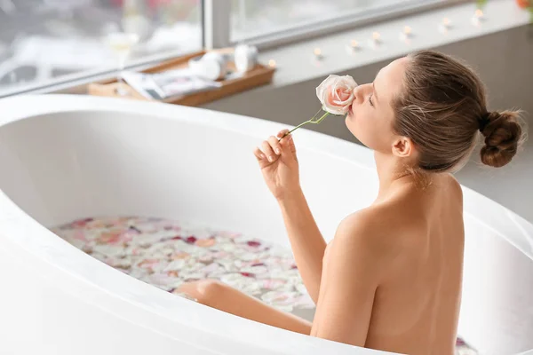 Piękna młoda kobieta bierze kąpiel z płatkami kwiatów w domu — Zdjęcie stockowe