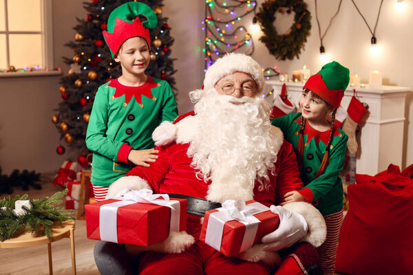 Santa Claus and little elf kids with gifts in room decorated for Christmas