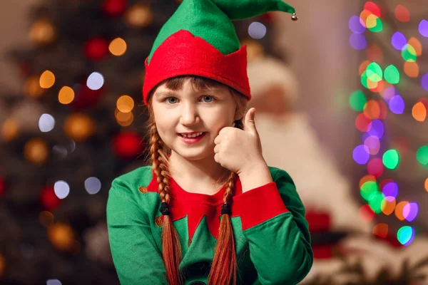 Little girl in costume of elf showing thumb-up gesture in room decorated for Christmas — Stock Photo, Image