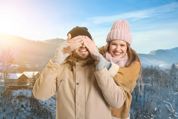 Lycklig kvinna som täcker ögonen på sin pojkvän nära bergsbyn på vinterdagen — Stockfoto
