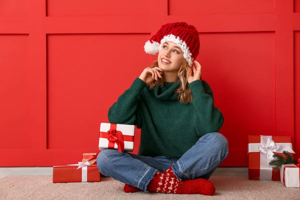 Happy young woman in winter clothes and with Christmas gifts sitting near color wall — Stock Photo, Image