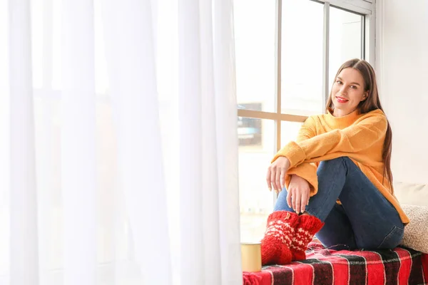 Junge Frau in Winterkleidung sitzt zu Hause auf Fensterbank — Stockfoto