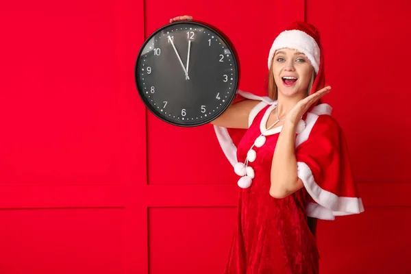 Giovane donna sorpresa in costume da Babbo Natale e con orologio sullo sfondo a colori. Conto alla rovescia di Natale — Foto Stock