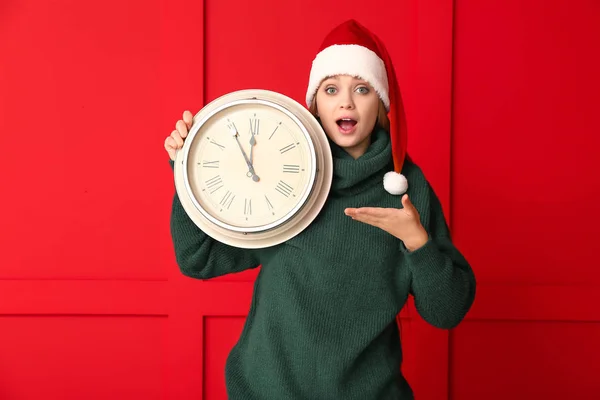Giovane donna sorpresa in cappello Babbo Natale e con orologio su sfondo a colori. Conto alla rovescia di Natale — Foto Stock
