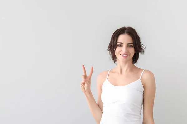 Beautiful young woman showing victory gesture on light background — Stock Photo, Image