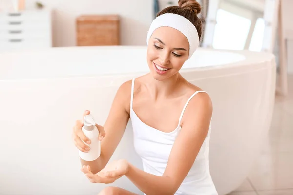 Beautiful young woman applying cream onto her skin in bathroom