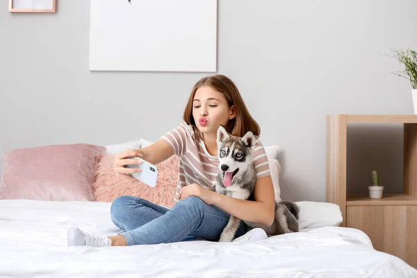 Cute teenage girl with funny husky puppy taking selfie on bed at home — Stock Photo, Image