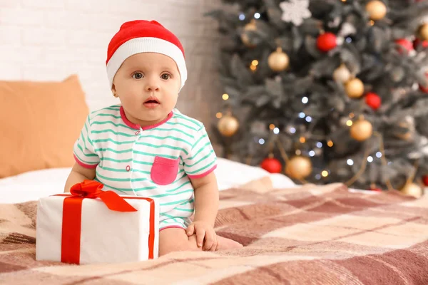 Niedliches kleines Baby mit Weihnachtsmann-Hut und Geschenk im weihnachtlich dekorierten Zimmer — Stockfoto