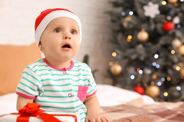 Cute little baby with Santa Claus hat and gift in room decorated for Christmas — Stock Photo, Image