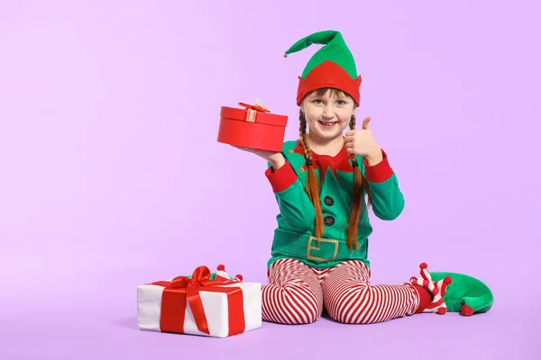 Menina em traje de elfo e com presentes mostrando gesto de polegar para cima no fundo de cor — Fotografia de Stock