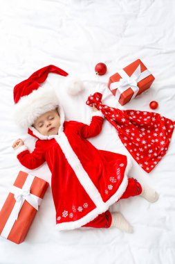 Noel Baba kostümü içinde küçük şirin bir bebek ve yatağında hediyeler var.