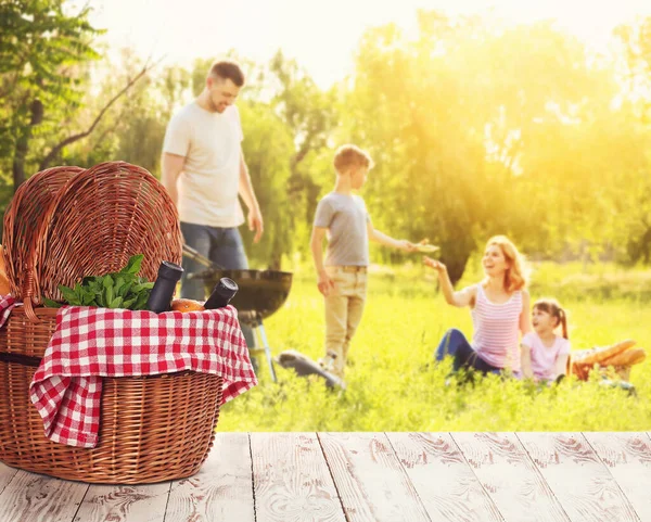 Wicker basket with tasty food and drink on table near happy family on summer picnic — Stock Photo, Image