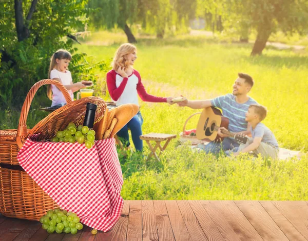 Cesta de vime com comida saborosa e bebida na mesa perto da família feliz no piquenique de verão — Fotografia de Stock